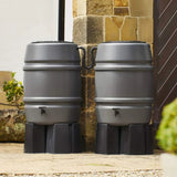 Two gray rain barrels with black stands against a stone wall.